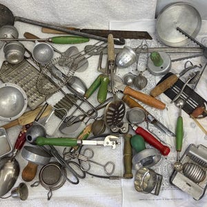 May include: A large collection of vintage metal kitchen utensils and tools, including graters, whisks, ladles, scoops, and various handheld gadgets with wooden and plastic handles in green, red, and natural wood tones scattered across a white surface.
