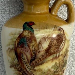 May include: A small ceramic jug with a cork stopper, featuring a glossy tan and cream finish. The front displays a detailed illustration of two pheasants in a natural landscape, with one bird showing vibrant green and red plumage on its head.