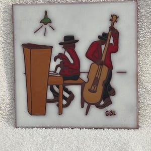 May include: A square decorative tile featuring a painted scene of a jazz duo. One figure plays a brown upright piano while wearing a red jacket and black hat. A second figure in a red jacket and black hat plays a large brown upright bass. GBL is signed.