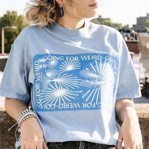 May include: A light blue t-shirt with a crew neck and short sleeves. The shirt features a rectangular blue graphic with white text and a starburst design. The text on the shirt reads "GOING FOR WEIRD" in a circular pattern.