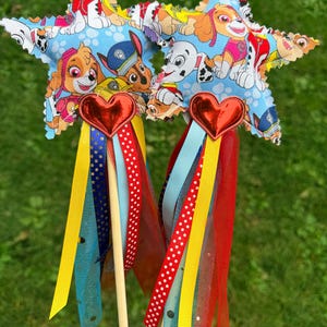 K&ouml;nnte beinhalten: Zwei sternf&ouml;rmige Zauberst&auml;be aus hellblauem Stoff mit Paw Patrol-Charakterdruck. Jeder Stern hat ein rotes Metallherz an der Basis, das an einem Holzstab befestigt ist. Eine Ansammlung bunter B&auml;nder in Gelb, Rot, Blau und mit Punkten h&auml;ngt am Herzen, zusammen mit Streifen aus rotem und blauem T&uuml;llstoff.