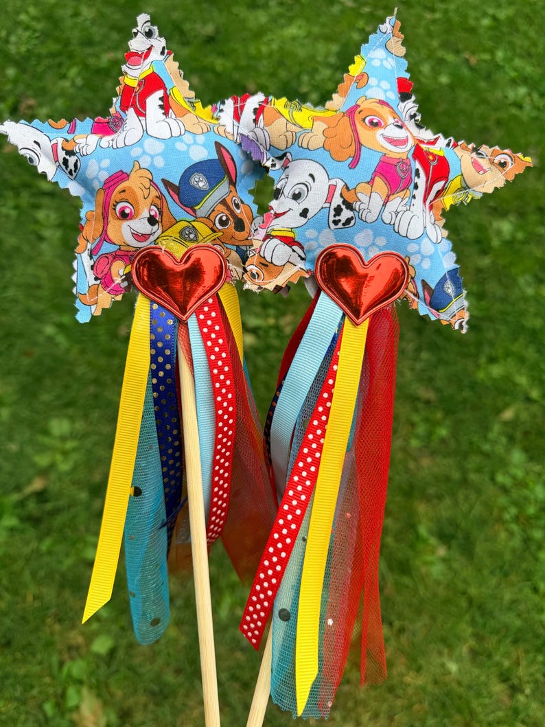 K&ouml;nnte beinhalten: Zwei sternf&ouml;rmige Zauberst&auml;be aus hellblauem Stoff mit Paw Patrol-Charakterdruck. Jeder Stern hat ein rotes Metallherz an der Basis, das an einem Holzstab befestigt ist. Eine Ansammlung bunter B&auml;nder in Gelb, Rot, Blau und mit Punkten h&auml;ngt am Herzen, zusammen mit Streifen aus rotem und blauem T&uuml;llstoff.
