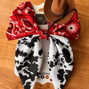 May include: A handmade hair bow featuring a red bandana print, a cow print, and a miniature brown cowboy hat. The bow is attached to a white card with the text "llaZima" and social media icons.