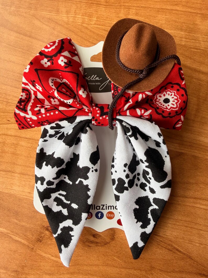 May include: A handmade hair bow featuring a red bandana print, a cow print, and a miniature brown cowboy hat. The bow is attached to a white card with the text "llaZima" and social media icons.