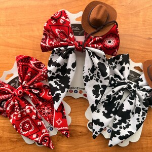 May include: Three hair bows with a western theme. One bow is red with a white paisley pattern, another is white with black cow spots, and the third is red with a white paisley pattern. Each bow has a small brown cowboy hat attached.
