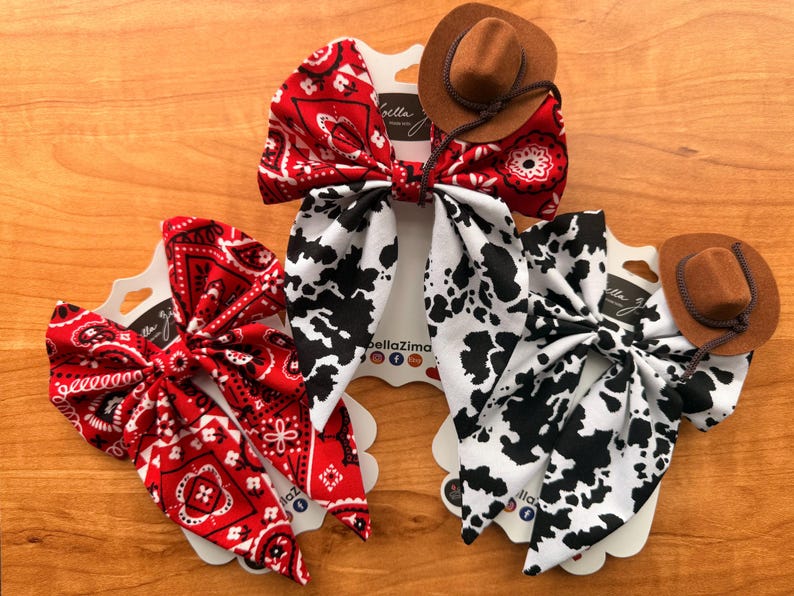 May include: Three hair bows with a western theme. One bow is red with a white paisley pattern, another is white with black cow spots, and the third is red with a white paisley pattern. Each bow has a small brown cowboy hat attached.
