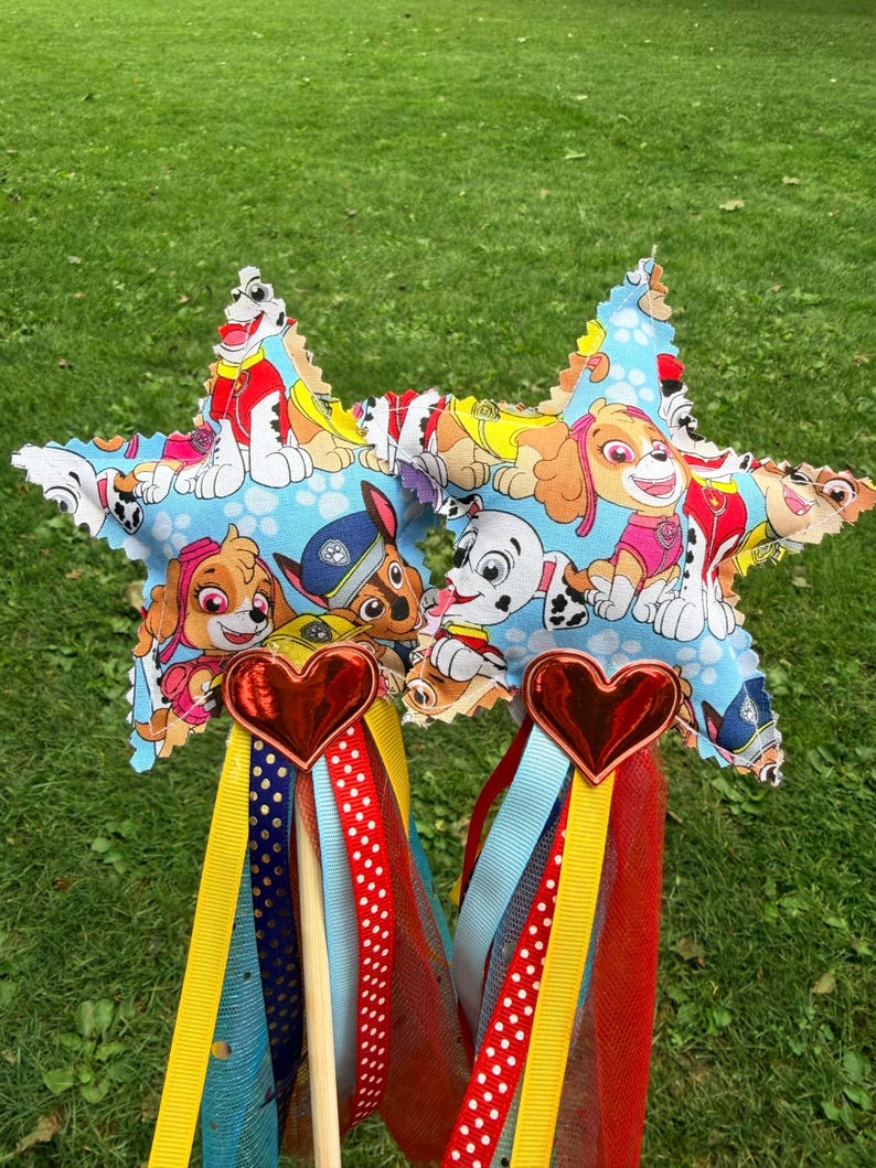 K&ouml;nnte beinhalten: Zwei sternf&ouml;rmige Zauberst&auml;be aus hellblauem Stoff mit Paw Patrol-Charakterdruck. Jeder Stab hat ein gl&auml;nzendes rotes Herz an der Basis des Sterns, befestigt an einem Holzstab mit herabh&auml;ngenden gelben, roten und blauen B&auml;ndern.