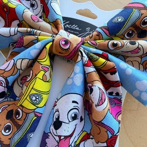K&ouml;nnte beinhalten: Eine Haarschleife aus Stoff mit einem bunten Muster aus Paw Patrol Zeichentrickwelpen auf hellblauem Grund. Die Schleife ist auf einer wei&szlig;en Pr&auml;sentationskarte befestigt, auf der oben mittig der Text Noella Zi in schwarzer Schrift steht.