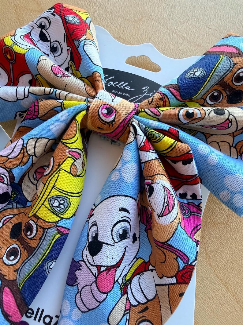 K&ouml;nnte beinhalten: Eine Haarschleife aus Stoff mit einem bunten Muster aus Paw Patrol Zeichentrickwelpen auf hellblauem Grund. Die Schleife ist auf einer wei&szlig;en Pr&auml;sentationskarte befestigt, auf der oben mittig der Text Noella Zi in schwarzer Schrift steht.