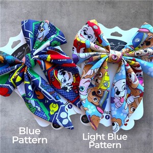 K&ouml;nnte beinhalten: Zwei Haarschleifen aus Stoff auf wei&szlig;en Pr&auml;sentationskarten. Die linke Schleife zeigt ein dunkelblaues Muster mit Paw Patrol-Figuren und geometrischen Formen. Die rechte Schleife hat ein hellblaues Muster mit verschiedenen Paw Patrol-Gesichtern.