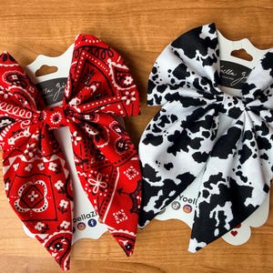 May include: Two fabric hair bows. One is red with a white paisley pattern. The other is white with black cow spots. Both are attached to white cardstock with the brand name "bella zi" printed on them.