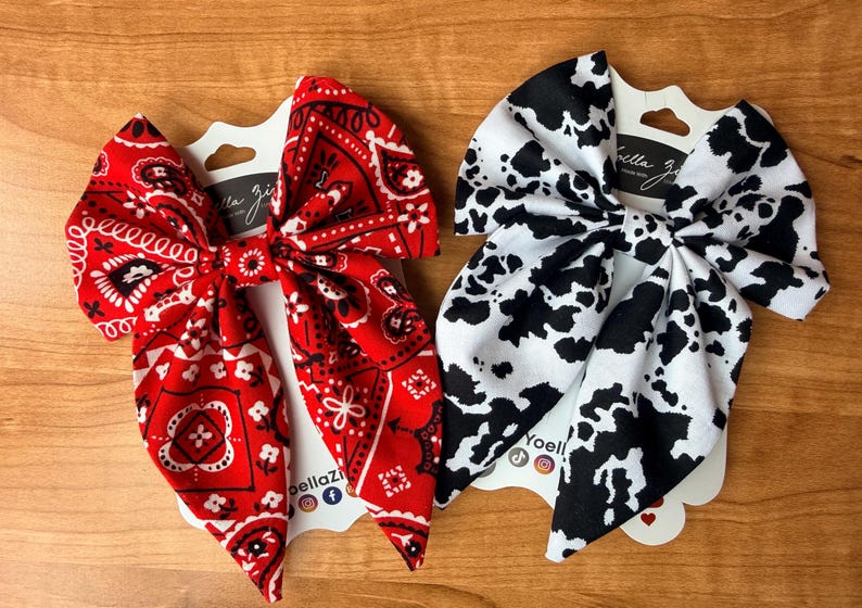 May include: Two fabric hair bows. One is red with a white paisley pattern. The other is white with black cow spots. Both are attached to white cardstock with the brand name "bella zi" printed on them.