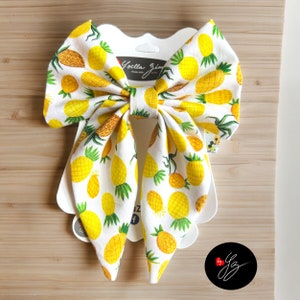 May include: A white bow with a pineapple print. The bow is tied with a loop and has two long tails. The bow is on a white background with the text "Yoella Jim" in black.