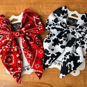 May include: Two fabric hair bows. One is red with a white paisley pattern, and the other is white with black cow spots. Each bow is attached to a white card with the brand name "bella zia" printed on it.