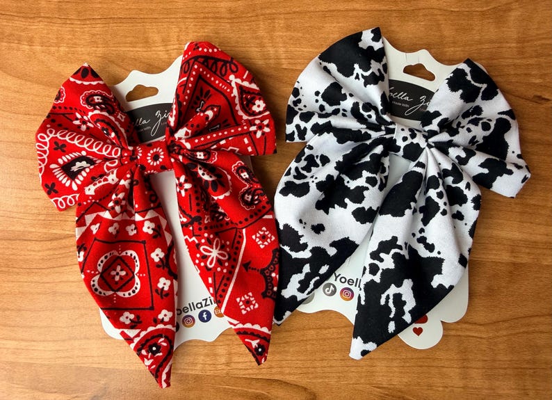 May include: Two fabric hair bows. One is red with a white paisley pattern, and the other is white with black cow spots. Each bow is attached to a white card with the brand name "bella zia" printed on it.