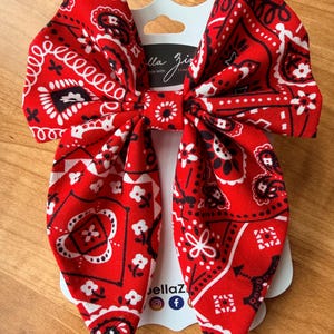 May include: A large red bandana bow with a white and black paisley pattern. The bow is attached to a white card with the text "Bella Ziz" and social media icons. The bow is made of fabric and is suitable for hair accessories.