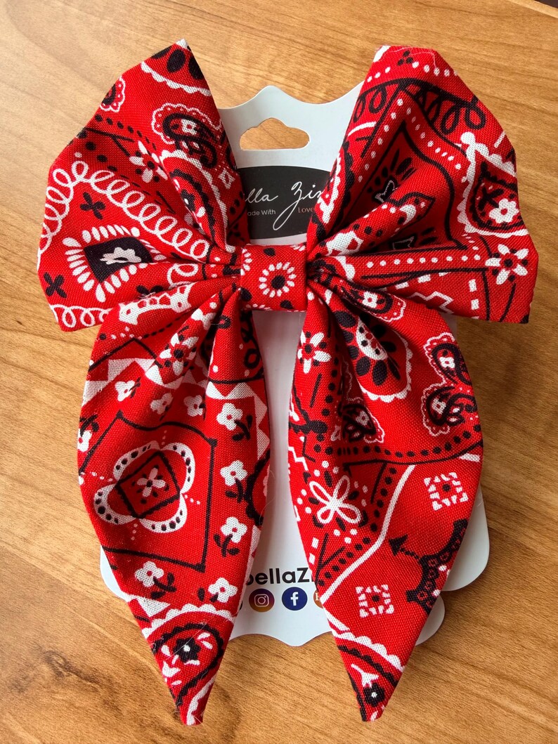 May include: A large red bandana bow with a white and black paisley pattern. The bow is attached to a white card with the text "Bella Ziz" and social media icons. The bow is made of fabric and is suitable for hair accessories.