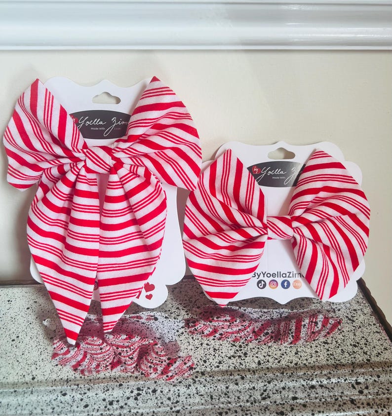 May include: Two red and white striped hair bows and several smaller matching bows are displayed on white cardstock. The bows are arranged on a textured surface. The bows are labeled with the text "by Yoella Zim".