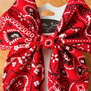 May include: A large red bandana bow with a white and black paisley pattern. The bow is attached to a white card with the text "ella zim" and "Made with Love".