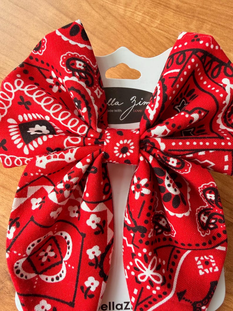 May include: A large red bandana bow with a white and black paisley pattern. The bow is attached to a white card with the text "ella zim" and "Made with Love".