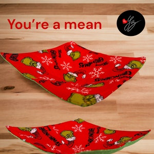 May include: Two red fabric bowls with a green lining. The bowls are decorated with a pattern of the Grinch, snowflakes, and the words "Merry Grinchmas". The text "You're a mean" is displayed in red.