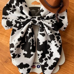 May include: A large hair bow with a black and white cow print pattern, paired with a miniature brown cowboy hat. The bow is attached to a white tag with the text "ella z" and social media icons.
