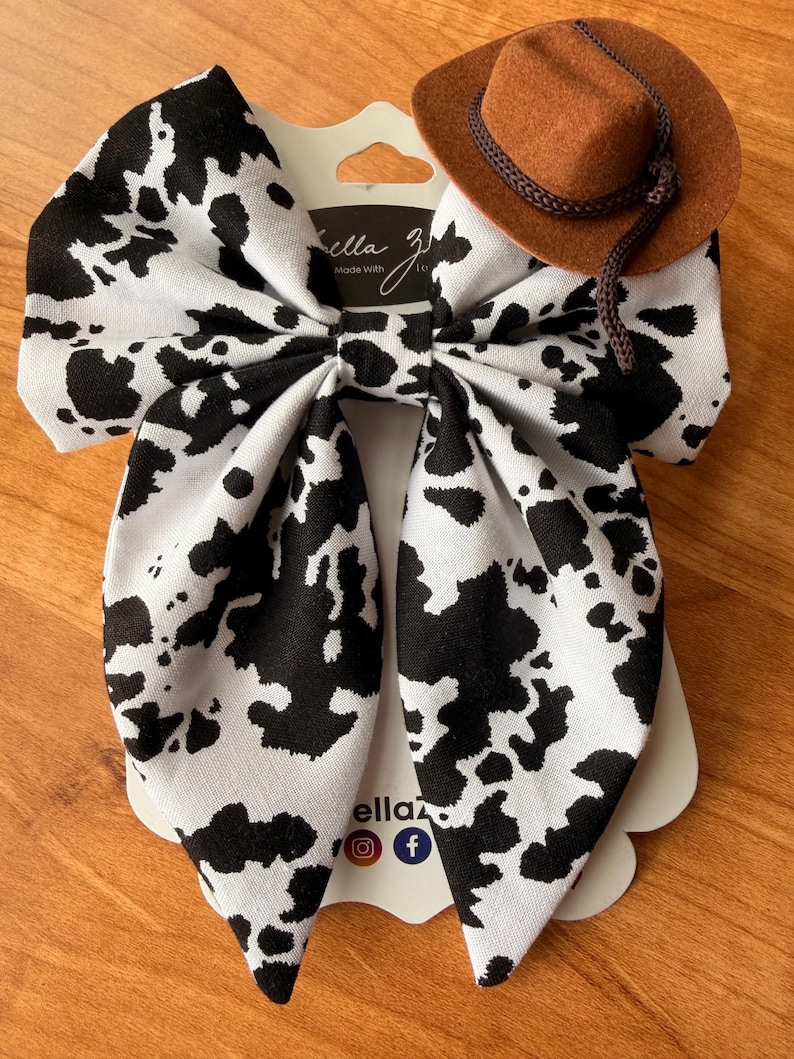 May include: A large hair bow with a black and white cow print pattern, paired with a miniature brown cowboy hat. The bow is attached to a white tag with the text "ella z" and social media icons.