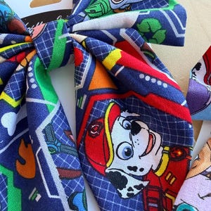 K&ouml;nnte beinhalten: Eine Haarschleife aus Stoff mit einem wiederkehrenden Muster aus Paw Patrol-Figuren, darunter der Dalmatiner Marshall mit rotem Helm, auf dunkelblauem Grund mit wei&szlig;em Gitter, gr&uuml;nen Recycling-Symbolen und gelben Schraubenschl&uuml;sseln.