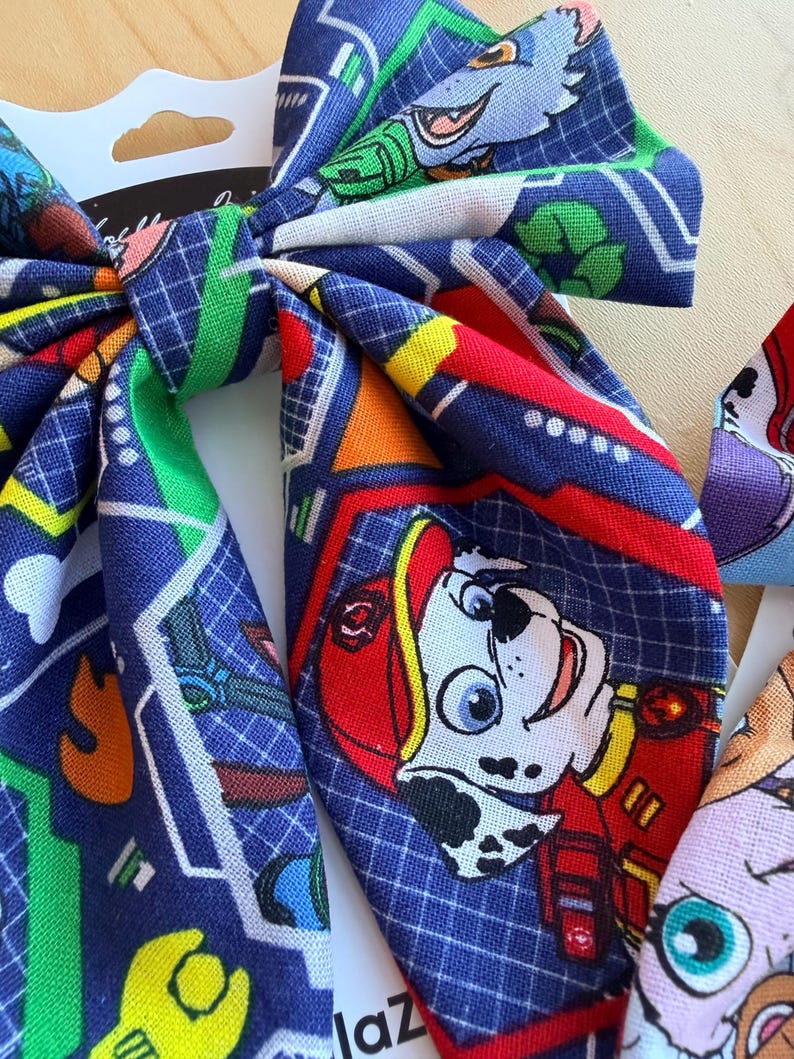 K&ouml;nnte beinhalten: Eine Haarschleife aus Stoff mit einem wiederkehrenden Muster aus Paw Patrol-Figuren, darunter der Dalmatiner Marshall mit rotem Helm, auf dunkelblauem Grund mit wei&szlig;em Gitter, gr&uuml;nen Recycling-Symbolen und gelben Schraubenschl&uuml;sseln.