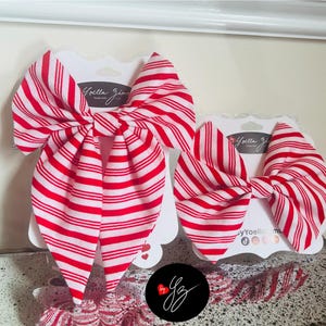 May include: Two red and white striped hair bows on white cardstock. The bows are in a classic bow shape, with a large bow and a smaller bow. The cardstock has the text "Yoella Zima" printed on it.
