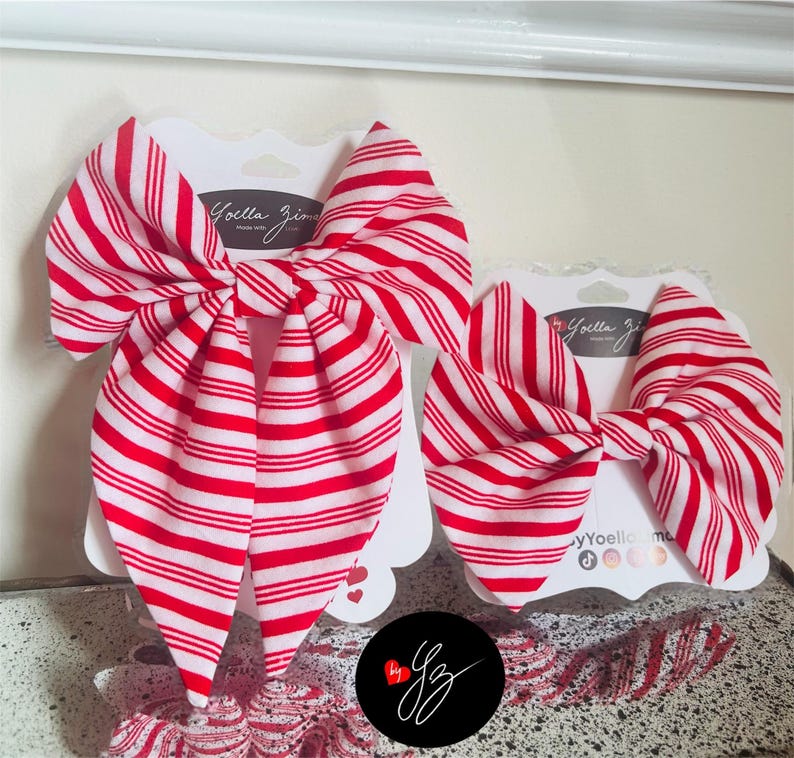 May include: Two red and white striped hair bows on white cardstock. The bows are in a classic bow shape, with a large bow and a smaller bow. The cardstock has the text "Yoella Zima" printed on it.