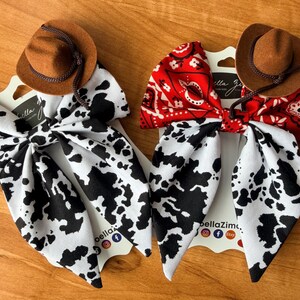 May include: Two hair bows with a Western theme. One bow features a black and white cow print, and the other has a red bandana pattern. Each bow is topped with a small brown cowboy hat. The bows are displayed on white cards with the text "bellaZim".