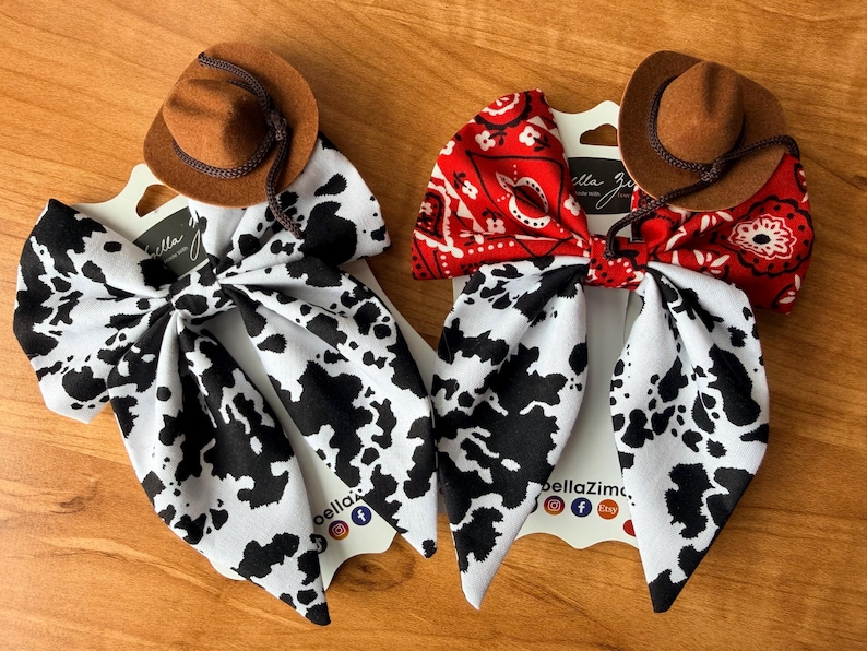 May include: Two hair bows with a Western theme. One bow features a black and white cow print, and the other has a red bandana pattern. Each bow is topped with a small brown cowboy hat. The bows are displayed on white cards with the text "bellaZim".