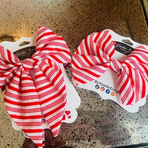 May include: Two red and white striped hair bows on white cardstock. The bows are tied in a large bow shape. The cardstock has the brand name "Yoella Zima" printed on it.