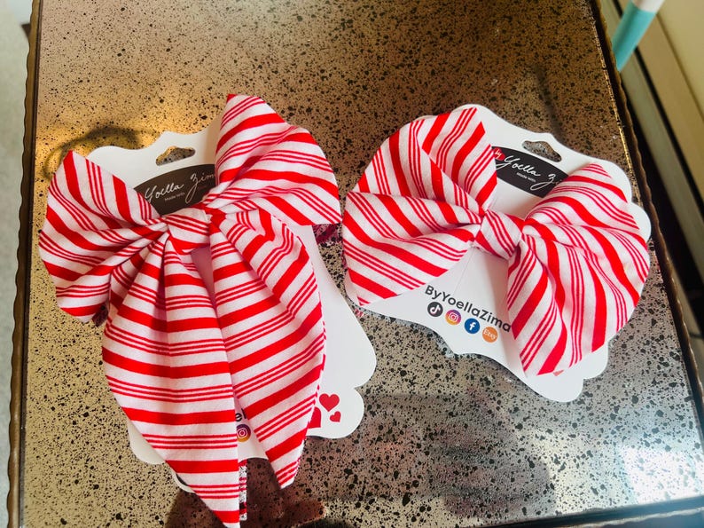 May include: Two red and white striped hair bows on white cardstock. The bows are tied in a large bow shape. The cardstock has the brand name "Yoella Zima" printed on it.