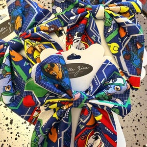 K&ouml;nnte beinhalten: Drei Haarspangen aus Stoff mit einem blauen, gr&uuml;nen, roten und gelben Cartoon-Figuren-Druck auf kariertem Hintergrund. Jede Schleife ist auf einer wei&szlig;en Karte mit dem in schwarzer Schrift gedruckten Text "Bella Zima" befestigt. Mehrlagiges Design.