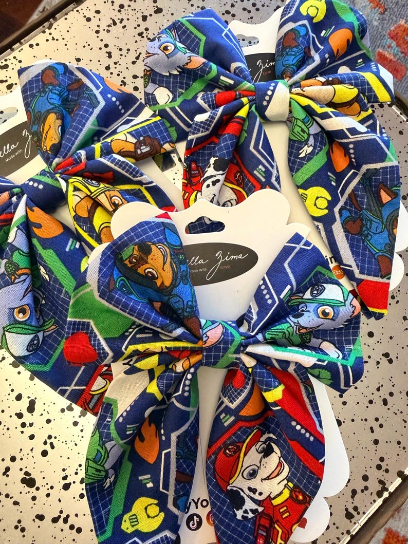 K&ouml;nnte beinhalten: Drei Haarspangen aus Stoff mit einem blauen, gr&uuml;nen, roten und gelben Cartoon-Figuren-Druck auf kariertem Hintergrund. Jede Schleife ist auf einer wei&szlig;en Karte mit dem in schwarzer Schrift gedruckten Text "Bella Zima" befestigt. Mehrlagiges Design.