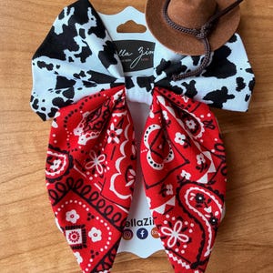 May include: A handmade hair bow with a cowboy theme. The bow features a cow print, red bandana print, and a small brown cowboy hat. The bow is attached to a white card with the text "ella Zim" and social media icons.