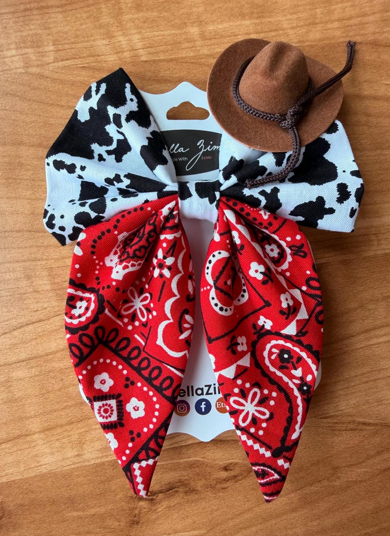 May include: A handmade hair bow with a cowboy theme. The bow features a cow print, red bandana print, and a small brown cowboy hat. The bow is attached to a white card with the text "ella Zim" and social media icons.