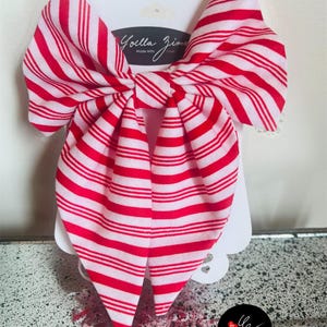 May include: A large red and white striped bow hair accessory. The bow is attached to a white card with the text "Joella Zima Made With Love". The bow is made of fabric and has a classic bow shape.