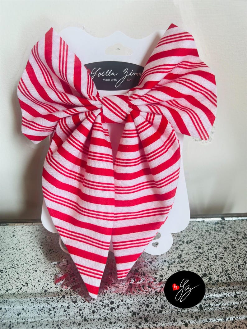 May include: A large red and white striped bow hair accessory. The bow is attached to a white card with the text "Joella Zima Made With Love". The bow is made of fabric and has a classic bow shape.