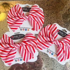 May include: Three red and white striped fabric bows on white cardstock tags. Each bow is tied in a classic bow shape. The tags have the text "ByYoellaZima" and social media icons. The bows are displayed on a gold surface.