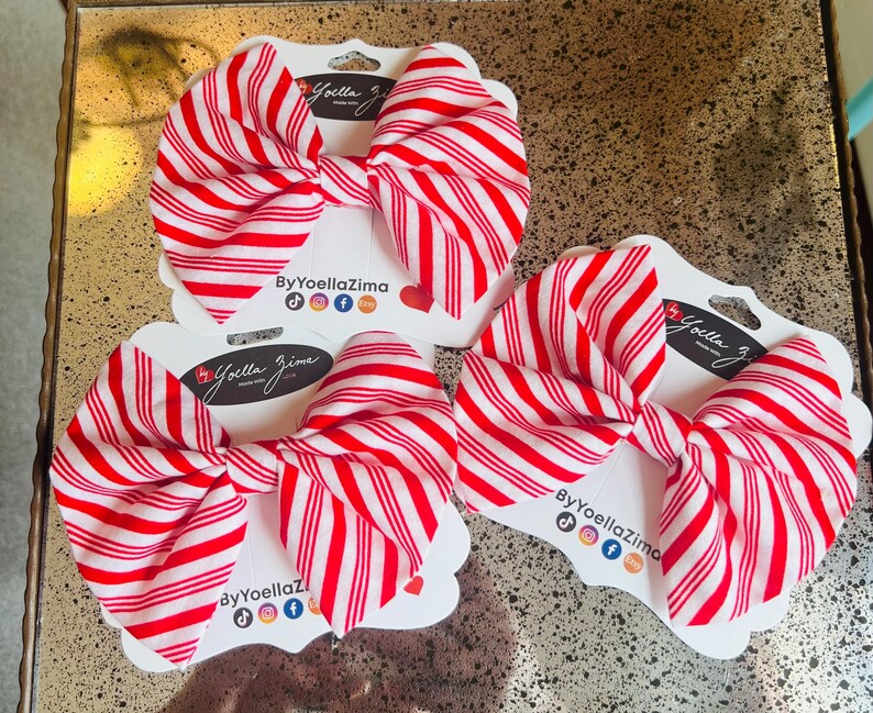 May include: Three red and white striped fabric bows on white cardstock tags. Each bow is tied in a classic bow shape. The tags have the text "ByYoellaZima" and social media icons. The bows are displayed on a gold surface.