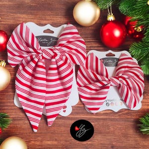 May include: Two red and white striped fabric bows on white cardstock, with the text "Yoella Zi". The bows are displayed on a wooden surface, with Christmas ornaments and greenery in the background. A black circle with a signature is in the bottom.