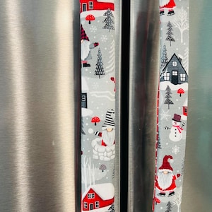 May include: Two gray and white fabric refrigerator handle covers with a winter gnome and house theme. The covers feature red houses, snowmen, gnomes, and trees.