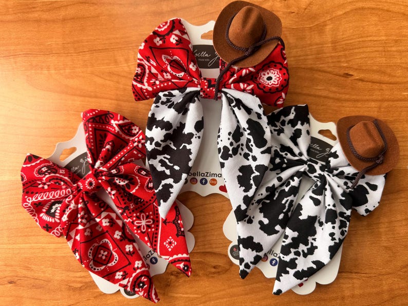 May include: Three hair bows with a Western theme. One bow is red with a white paisley pattern, another is black and white cow print, and the third is red paisley. Each bow has a small brown cowboy hat attached.