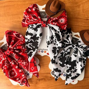 May include: Three hair bows with a Western theme. Two bows are red with white paisley patterns, and one is white with black cow spots. Each bow is topped with a small brown cowboy hat.