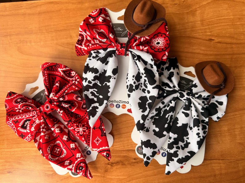 May include: Three hair bows with a Western theme. Two bows are red with white paisley patterns, and one is white with black cow spots. Each bow is topped with a small brown cowboy hat.