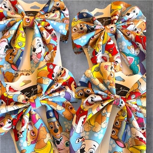 K&ouml;nnte beinhalten: Vier Haarschleifen mit einem sich wiederholenden Muster aus Paw Patrol Zeichentrickhunden auf hellblauem Grund. Jede Schleife ist auf einer wei&szlig;en Karte mit dem in Schwarz gedruckten Text Yoella Zima befestigt. Der Stoff hat eine glatte Oberfl&auml;che.
