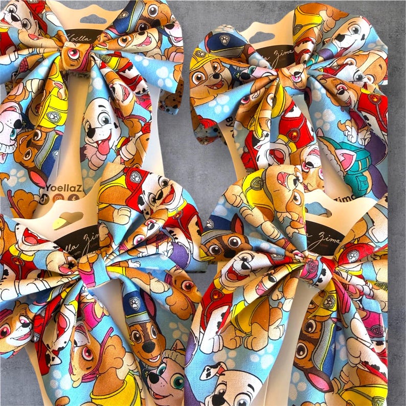 K&ouml;nnte beinhalten: Vier Haarschleifen mit einem sich wiederholenden Muster aus Paw Patrol Zeichentrickhunden auf hellblauem Grund. Jede Schleife ist auf einer wei&szlig;en Karte mit dem in Schwarz gedruckten Text Yoella Zima befestigt. Der Stoff hat eine glatte Oberfl&auml;che.